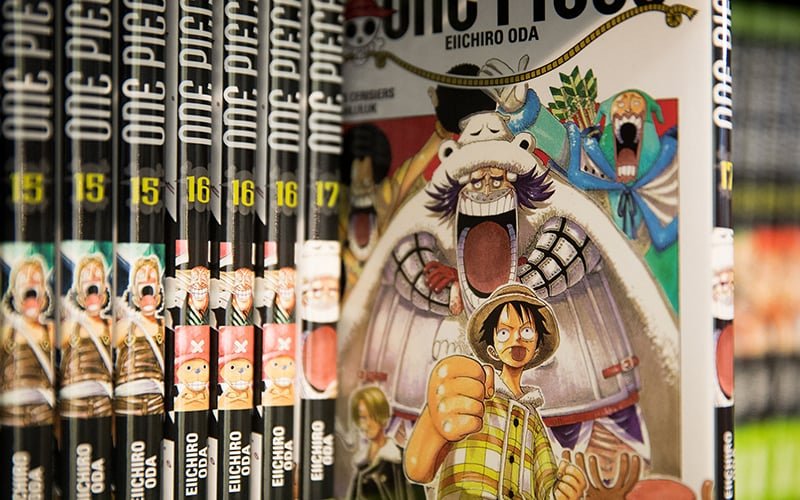 An open laptop displaying One Piece manga online, surrounded by books and coffee in a cozy reading setting.